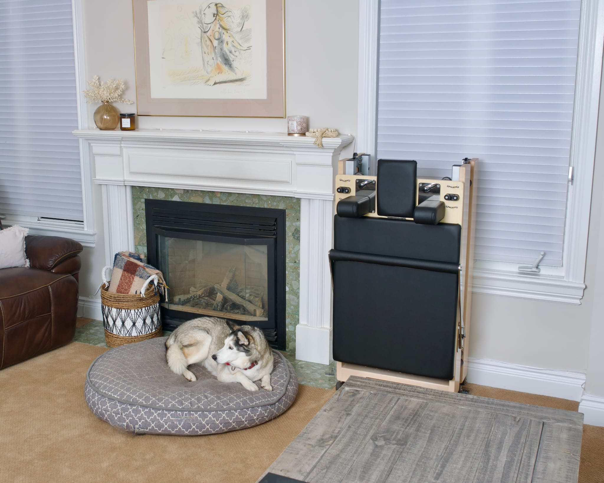 A folded Maple Elite Home Folding Reformer stored in a living room next to a fire place.